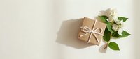 Kraft paper gift box tied with white bow beside fresh white flowers on beige background with soft shadows, top view flat lay.