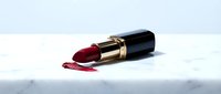 Luxury burgundy lipstick with gold trim and black cap on white marble surface with smudged color swatch for cosmetics, beauty, and makeup design projects.