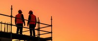 Two construction workers in hard hats standing on scaffolding silhouetted against vibrant orange sunset sky for industrial safety and teamwork concepts.