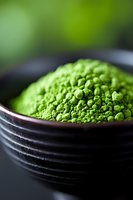 Vibrant green matcha tea powder in black ceramic bowl with soft focus background, macro shot showing fine texture and natural organic appearance.