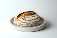 Fresh artisanal sourdough bread loaf with crispy crust and flour dusting on white ceramic plate against neutral background, minimalist food photography.