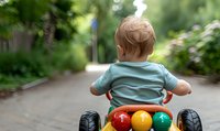 Toddler boy in light blue t-shirt riding colorful plastic toy car on garden path, rear view with blurred natural background showing summer outdoor activity.
