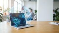 Modern laptop displaying hexagonal molecular structure on screen in bright office workspace with blurred professionals working in background.