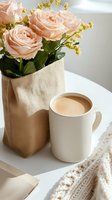 Soft pink roses and yellow wildflowers in beige textile bag next to white coffee mug on table with knitted throw blanket, creating cozy morning atmosphere.