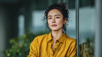 Young Asian businesswoman in yellow shirt standing confidently in modern office environment with natural lighting and professional corporate atmosphere.