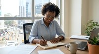 Young African American businesswoman writing notes at office desk with laptop and coffee cup in modern workplace with city view.