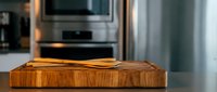 Wooden cutting board on kitchen counter with modern stainless steel appliances in background. Natural wood grain texture for culinary preparation and cooking.