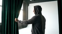 Young man in dark hoodie opening green curtains by window in modern interior room with natural daylight streaming through glass.