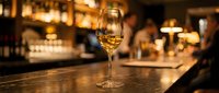 Glass of white wine on wooden bar counter in upscale restaurant with warm ambient lighting and blurred background creating elegant dining atmosphere.