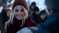 Young blonde woman smiling outdoors on winter evening, wearing burgundy knit beanie and scarf, with blurred crowd and bokeh lights in background.
