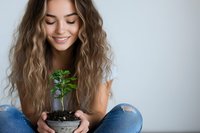 Young Asian woman with long wavy hair holding small plant in ceramic pot, sitting cross-legged in casual ripped jeans against light wall background, expressing joy and care.