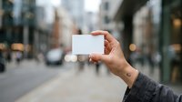 Close-up of hand holding blank white business card on urban street with blurred city background and pedestrians for branding mockup.