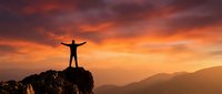 Silhouette of person with raised arms celebrating success on mountain peak at dramatic sunset with orange and purple sky over mountain ranges.
