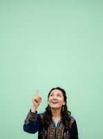 Young woman with long dark hair and East Asian ethnicity wearing an ornate navy blue embroidered jacket, pointing upward with index finger while smiling and looking up against mint green background.