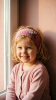Adorable young girl with curly blonde hair wearing pink knitted headband and cardigan smiling by window. Cozy childhood portrait for family lifestyle content.