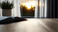 Cozy reading corner with open book and potted plant on wooden table against window with sunset view and soft curtains, creating peaceful atmosphere.