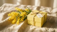 Yellow gift box with twine bow and mimosa flowers on linen fabric in warm sunlight for spring celebration and International Women's Day greeting card design.