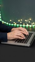 Hands typing on a laptop with festive green and gold bokeh lights in the background, capturing holiday remote work and online shopping season.
