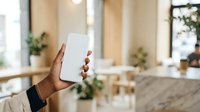 Person with dark skin tone holding blank white smartphone in modern office or home interior with plants and natural lighting for app mockup or technology presentation.