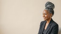 Middle-aged Black woman with gray braided updo hairstyle wearing dark blazer, smiling while looking to the side against neutral beige background. Corporate professional portrait.