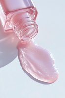 Pink glossy liquid spilling from glass bottle neck creates shimmering drop on white surface, highlighting luxurious cosmetic texture and transparency.