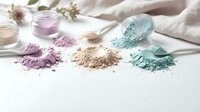Natural cosmetic clay powders in pastel colors scattered on white surface with flowers and fabric for spa beauty treatments and skincare routines.