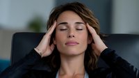 Young woman with closed eyes massaging temples, experiencing headache or stress while sitting on black leather couch in modern living room.