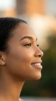 Young African American woman smiling in warm golden hour sunlight with natural makeup and earrings showing confidence and happiness in outdoor portrait.