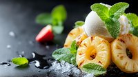 Grilled pineapple rings with vanilla ice cream scoop and fresh mint leaves on dark background with water drops, served with strawberry slice.