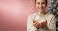 Young woman holding glowing Christmas ornament with warm lights against pink background near decorated tree for holiday celebration and festive season greeting cards.