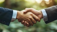 Business handshake between two male executives in suits outdoors with natural bokeh background, representing partnership, agreement and trust.