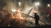 Emergency responder firefighter in protective gear battling intense aircraft crash scene with dramatic flames and smoke at night under bright floodlights.