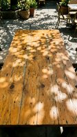 Wooden outdoor dining table with dappled sunlight casting leaf shadows across weathered planks in a garden patio setting with potted plants and chairs.