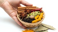 Hand holding wicker basket filled with aromatic spices including cinnamon sticks, star anise, cardamom, turmeric and bay leaves for cooking.