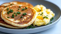 Fluffy homemade pancakes served on gray plate with scrambled eggs garnished with fresh chopped chives, breakfast close up with selective focus and natural lighting.