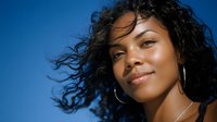 Young African American woman with curly black hair smiling against blue sky background, wearing silver hoop earrings. Natural beauty and confidence portrait for lifestyle and fashion content.