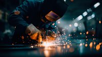 Industrial welder in protective mask working with metal, creating bright orange sparks in dark workshop environment.