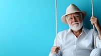 Senior Caucasian man with white beard wearing panama hat and white shirt sitting on rope swing against bright blue background for summer leisure concept.