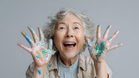 Senior woman with colorful paint on hands expressing joy and playfulness against gray background, showcasing creativity and active aging.