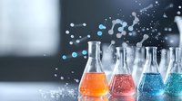 Chemical laboratory glassware with colorful liquid solutions in orange, pink and blue creating dynamic splash effect against blurred background.
