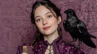 Young woman with black raven bird perched on shoulder against ornate purple wallpaper background for fantasy portrait photography and storytelling concepts.