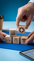 Male hands arranging wooden blocks with healthcare icons including stethoscope, heart, medical cross, and caduceus symbol on blue background for insurance and wellness concepts.