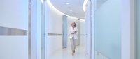 Middle-aged woman with light skin tone, wearing professional business attire, walking through modern office corridor with glass partitions and contemporary lighting design.
