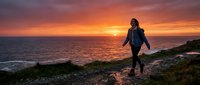 Woman in wetsuit walking toward ocean at golden sunset, ready for surfing or water sports adventure on coastal beach.