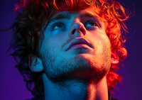 Young male model with curly hair in dramatic neon lighting against purple background, looking up with intense expression. Red and blue color contrast creates moody atmosphere.
