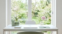 Minimalist white desk workspace by bright window with blooming garden view, laptop, notebook and pink flower in vase creating peaceful home office atmosphere.