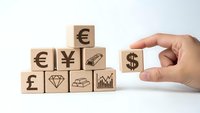 Hand placing wooden cube with dollar sign among blocks featuring currency symbols and financial icons on white background for investment concepts.