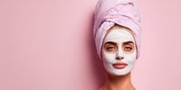 Young woman with facial mask and towel on head enjoying spa treatment on pink background for skincare and beauty routine.