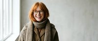 Smiling middle-aged Caucasian businesswoman with red hair and glasses wearing brown coat and scarf in modern office interior for professional portraits.