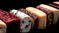 Assorted gourmet cheese and sausage slices arranged in diagonal row showing variety of textures and patterns, close-up view on dark background for food photography.